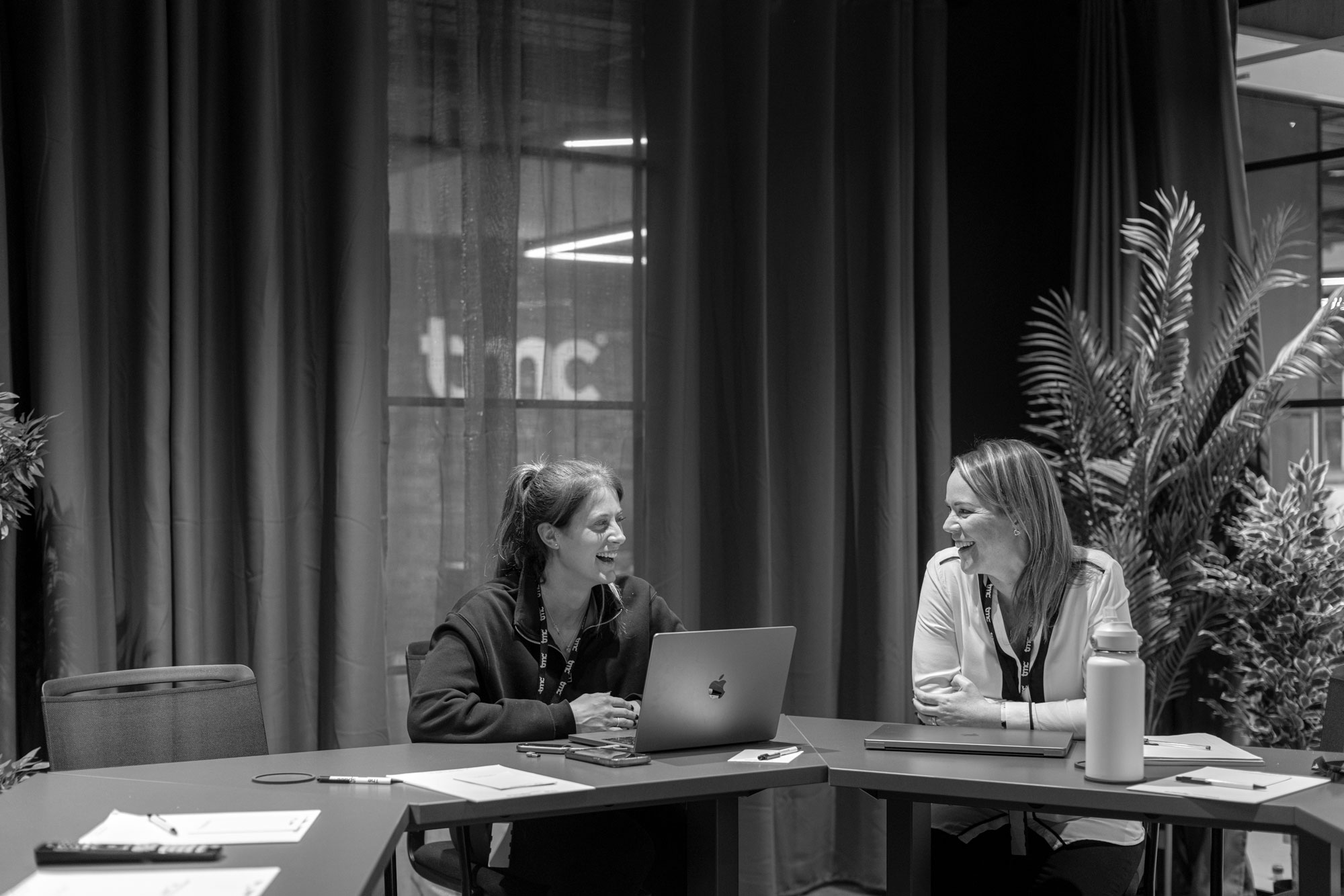 Two women are sitting in the boardroom, smiling at each other with a laptop open on the table